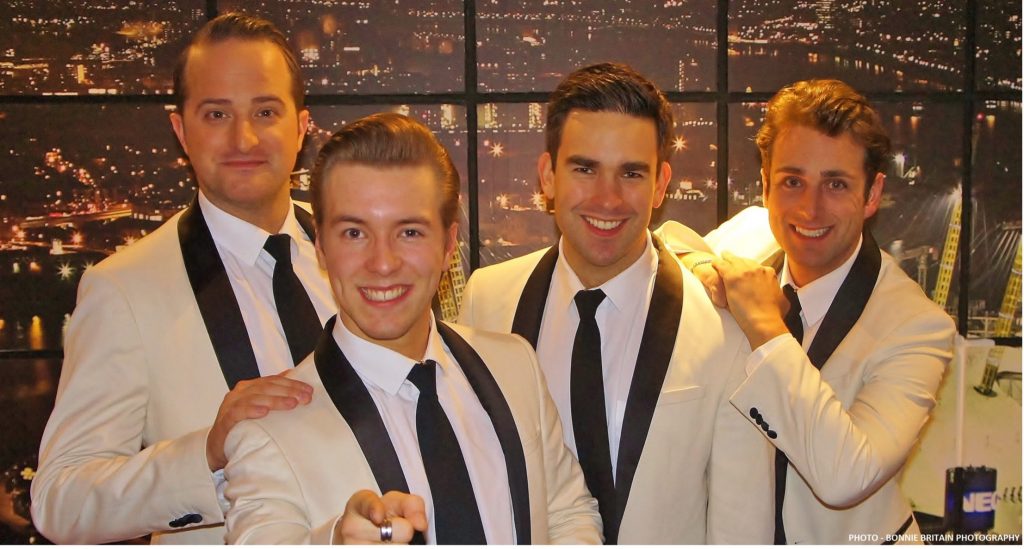 This is a photo of the 4 singers in white jackets posing in a green room